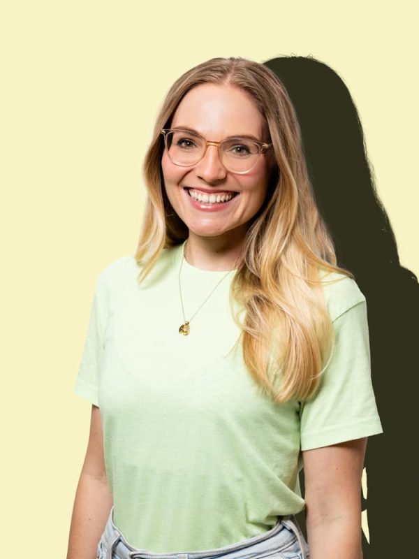 Frau mit langen blonden Haaren und Brille in hellgrünem Shirt vor gelbem Hintergrund